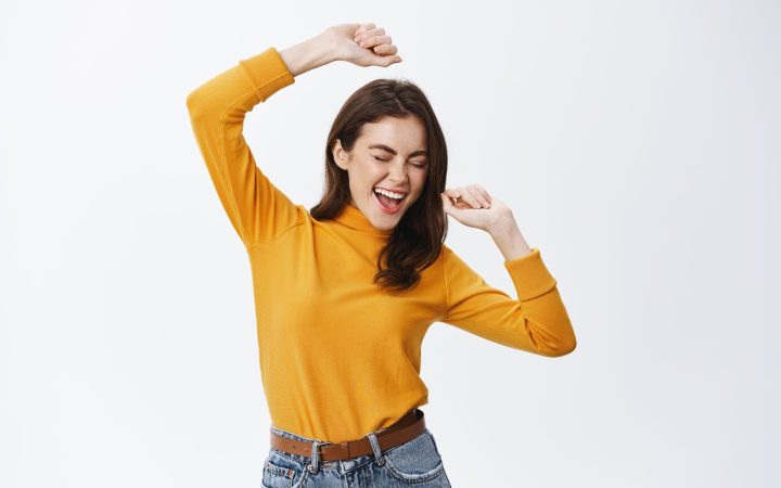 happy-attractive-woman-dancing-having-fun-raising-hands-up-carefree-enjoying-music-standing-against-white-wall