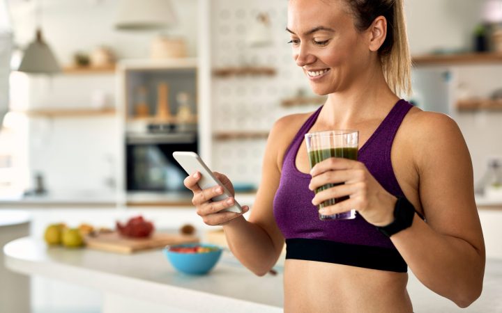 young-happy-sportswoman-using-smart-phone-while-drinking-detox-smoothie-kitchen